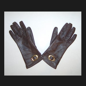 Könnte beinhalten: Ein Paar dunkelbraune Lederhandschuhe. Jeder Handschuh hat eine goldfarbene Schnalle am Handgelenk. Die Handschuhe sind vor einem weißen Hintergrund abgebildet.