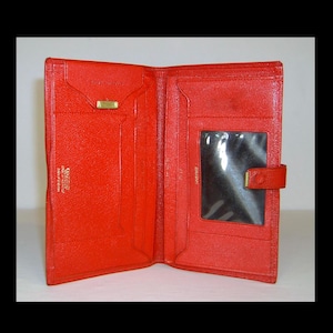 Puede incluir: Cartera de cuero roja abierta con múltiples compartimentos, incluyendo una ventana transparente para identificación y un cierre dorado. La cartera está abierta, mostrando su diseño interior y textura.