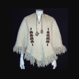 May include: Cream-coloured poncho with fringe trim and decorative embroidery. The poncho features a V-neckline with a tassel detail and colourful embroidered patterns on the front. The bottom edge has a fringe.