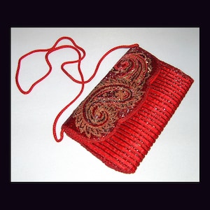 Puede incluir: Un clutch rojo con un diseño de cachemira decorativo en abalorios dorados y bronce. El clutch tiene una correa de cordón rojo a juego y una textura acanalada. Un accesorio vibrante.