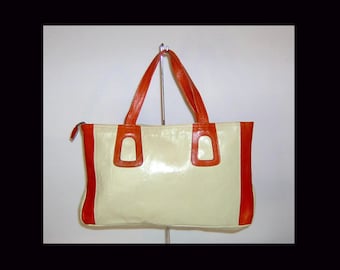 May include: A rectangular handbag featuring a cream-colored central panel and vibrant orange side panels. The bag includes two orange leather handles attached to the top with orange rectangular accents and a side zipper closure for the main compartment.