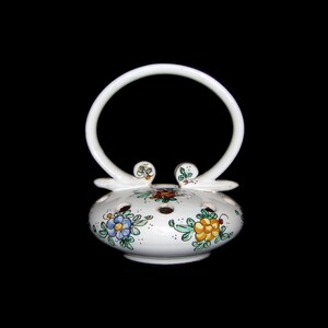 May include: A white ceramic basket with a circular handle and floral designs in blue, yellow, and green. The basket has small holes around the top and decorative accents near the handle's base. The background is black.