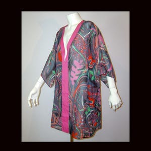 Puede incluir: Una túnica transparente con escote en V profundo y mangas anchas, con un estampado abstracto multicolor vibrante en tonos rosa, verde, rojo y gris oscuro. La prenda está acentuada con un borde vertical de color rosa sólido en la abertura frontal.