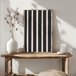 Puede incluir: Un cuadro enmarcado con un patrón de rayas verticales negras y blanco roto. La obra de arte se exhibe sobre una consola de madera, junto a un jarrón blanco con flores secas y otros elementos decorativos. La estética general es minimalista y moderna.