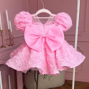 Peut inclure: Une robe rose avec un grand nœud et des détails floraux texturés. La robe présente des manches bouffantes et un décolleté transparent. Elle est suspendue à un cintre blanc.