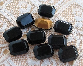 May include: A group of ten black, faceted, rectangular glass gems. One of the gems is gold.