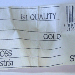 Pu&ograve; includere: Un'etichetta bianca con testo nero che recita "1012 SS 19 CRYSTAL 10/00/00 GROSS Made in Austria 1st QUALITY GOLD" e un codice a barre. L'etichetta presenta anche un logo Swarovski in bianco e nero con il testo "SWAROVSKI".