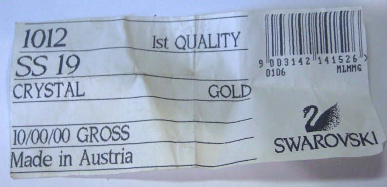 Pu&ograve; includere: Un'etichetta bianca con testo nero che recita "1012 SS 19 CRYSTAL 10/00/00 GROSS Made in Austria 1st QUALITY GOLD" e un codice a barre. L'etichetta presenta anche un logo Swarovski in bianco e nero con il testo "SWAROVSKI".