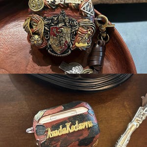 May include: Two Airpod cases, one with a red, gold, and black design with the words "Avada Kedavra" and the other with a red, gold, and brown design with the words "Gryffindor" and a Hogwarts crest.