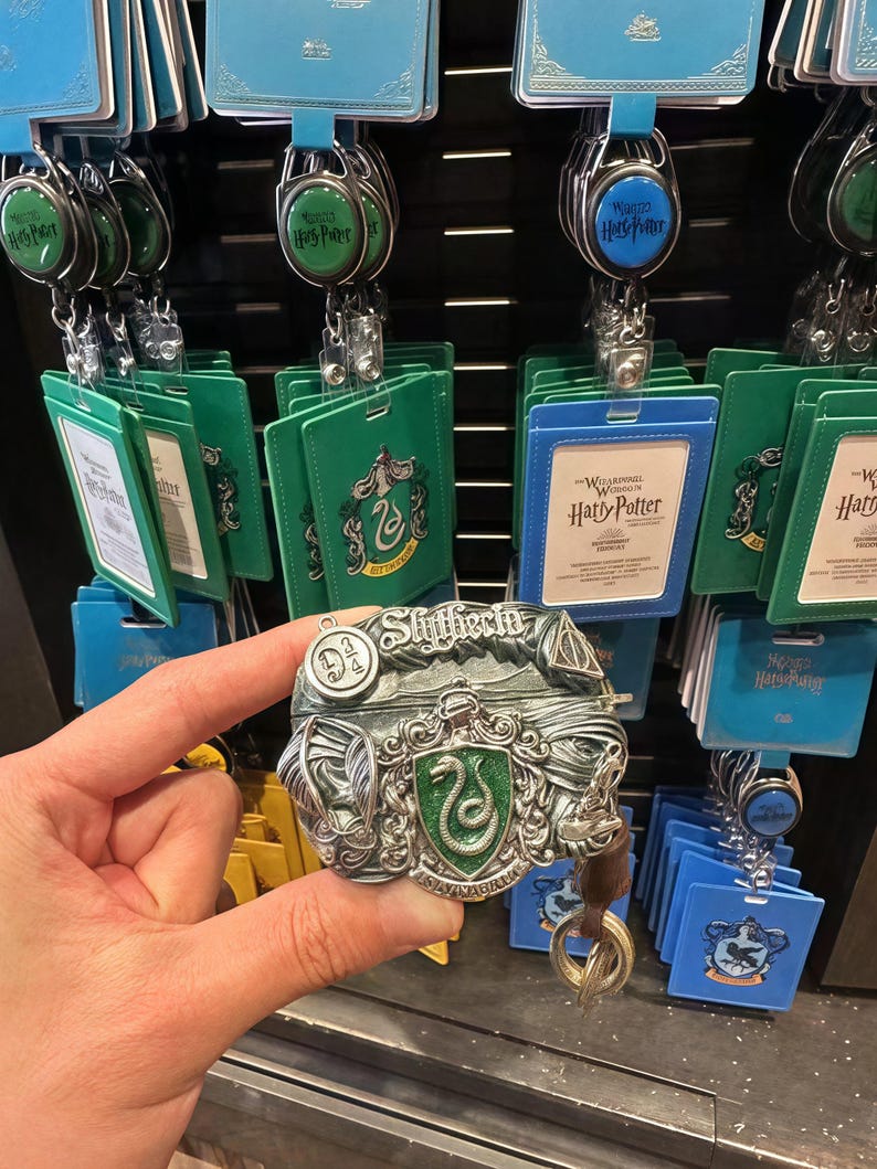 May include: A silver-colored Slytherin house crest keychain with a green snake and the words "Slytherin" and "Serpentes" on it. The keychain is attached to a brown leather strap with a metal ring.