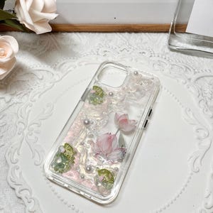 Pu&ograve; includere: Una custodia per telefono trasparente con un design floreale rosa e verde. Il design presenta fiori di loto, foglie e accenti perlati.