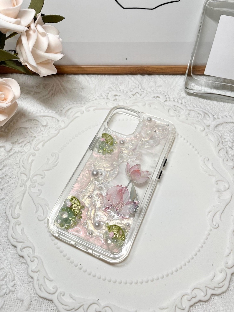 Pu&ograve; includere: Una custodia per telefono trasparente con un design floreale rosa e verde. Il design presenta fiori di loto, foglie e accenti perlati.