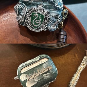 May include: A green and silver Airpod case with a Slytherin crest and the words "Slytherin" and "9 3/4" on it. The case is sitting on a wooden surface. A second Airpod case is green and white with the words "Amanda Kedavra" on it. The case is sitting on a wooden surface next to a silver pen.