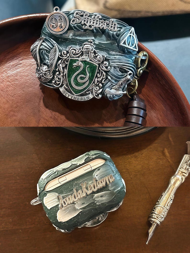 May include: A green and silver Airpod case with a Slytherin crest and the words "Slytherin" and "9 3/4" on it. The case is sitting on a wooden surface. A second Airpod case is green and white with the words "Amanda Kedavra" on it. The case is sitting on a wooden surface next to a silver pen.
