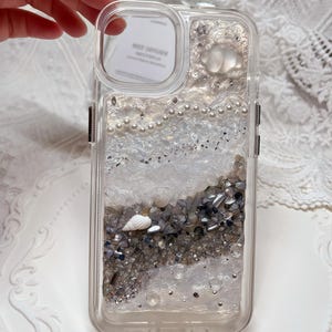 Può includere: Una custodia per telefono trasparente con un design a glitter bianco e argento. La custodia presenta una conchiglia, perle e piccole perline argentate.