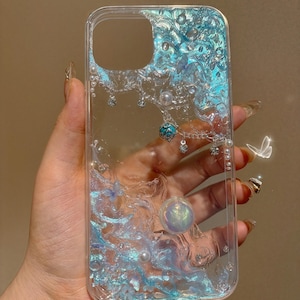 Puede incluir: Una funda transparente para teléfono con un líquido azul y purpurina en su interior. La funda tiene una perla y un dije turquesa colgando.