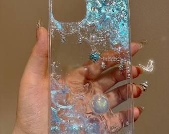 May include: A clear phone case with a blue liquid and glitter inside. The case has a pearl and a turquoise charm hanging from it.