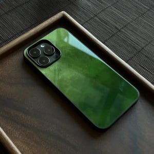 Könnte beinhalten: Eine grüne Handyhülle mit glänzendem Finish und einem Ausschnitt für eine Dreifach-Kamera. Die Hülle liegt auf einem Holztablett mit dunkler, strukturierter Oberfläche.