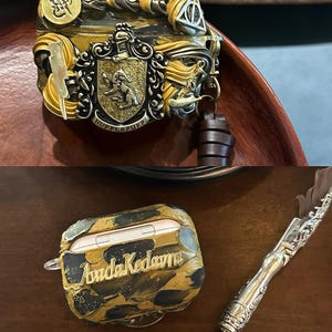 May include: A gold and black Airpod case with a marbled design and the name "Avada Kedavra" written in gold lettering. The case is sitting on a wooden surface next to a silver pen with a feather nib.