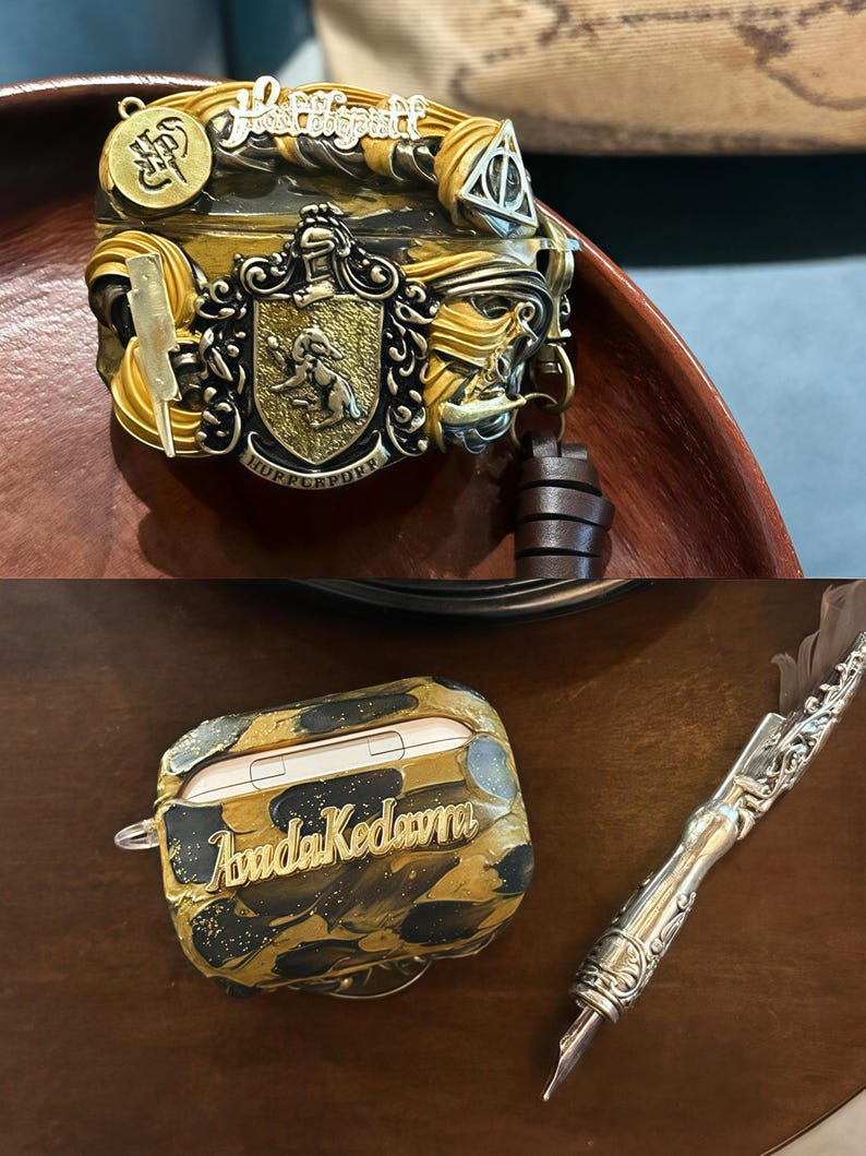 May include: A gold and black Airpod case with a marbled design and the name "Avada Kedavra" written in gold lettering. The case is sitting on a wooden surface next to a silver pen with a feather nib.