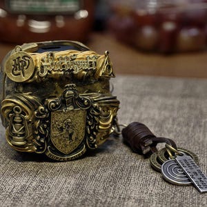 May include: A gold-colored metal charm with a Hogwarts crest and the words "Hufflepuff" and "I'll be there for you" on a leather strap. The charm is decorated with a crest and other details.