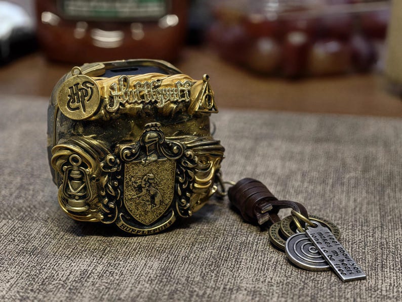 May include: A gold-colored metal charm with a Hogwarts crest and the words "Hufflepuff" and "I'll be there for you" on a leather strap. The charm is decorated with a crest and other details.