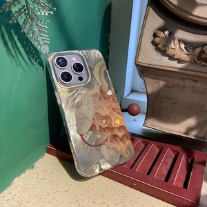 Puede incluir: Una funda de iPhone dorada con un diseño de serpiente y flores. La funda tiene un acabado holográfico y una protuberancia de cámara elevada.
