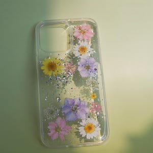 Puede incluir: Una funda transparente para teléfono con fondo brillante y flores prensadas en tonos rosa, morado, amarillo y blanco.