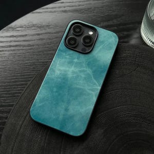 Könnte beinhalten: Türkis marmoriertes Handyhülle mit schwarzem Kameramodul. Die Hülle liegt auf einer dunklen Holzoberfläche. Die Handyhülle hat ein glänzendes Finish und ein schlankes Design.