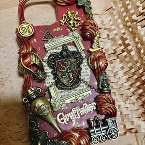 Może przedstawiać: Czerwono-złote etui na telefon z herbem Gryffindoru, złotym zniczem, pociągiem i butelką z eliksirem. Etui jest ozdobione złotem i ma herb Gryffindoru w środku.
