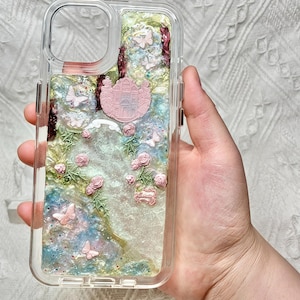 Op de afbeelding: Transparante telefoonhoes met een decoratief interieur met roze rozen, vlinders en een roze kasteelembleem. De hoes heeft een glinsterende, iriserende achtergrond met groene en blauwe tinten. De hoes wordt in een hand gehouden.