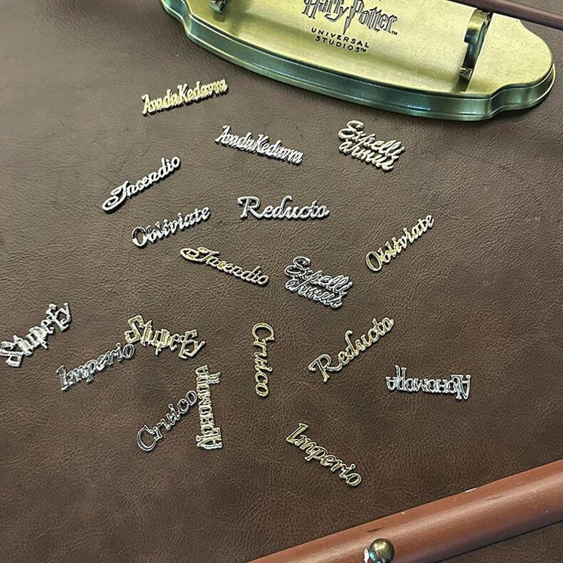 May include: A collection of gold and silver charms with various Harry Potter spells written on them, including "Avada Kedavra", "Reducto", "Imperio", "Stupefy", "Expelliarmus", "Obliviate", and "Facendio".