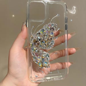 May include: A clear phone case with a sparkly butterfly design. The butterfly is made of rhinestones and is iridescent.