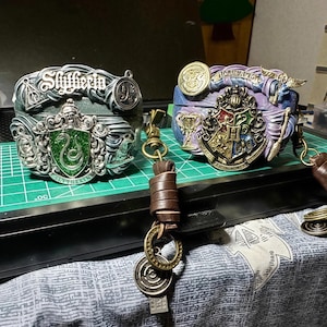 May include: Two silver and gold Harry Potter house crest keychains. The keychain on the left features the Slytherin crest with a green snake and the words "Slytherin" on the bottom. The keychain on the right features the Hogwarts crest with the words "Hogwarts School of Witchcraft and Wizardry" on the bottom. Both keychains have a brown leather strap.