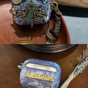 May include: A blue and gold Hogwarts themed phone case with a galaxy design and the name "Aurora Kedawra" written in gold lettering. The case is sitting on a wooden surface next to a silver pen.