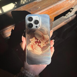 May include: A clear phone case with a gold and red design of two goldfish swimming in a circle. The case is designed for an iPhone.