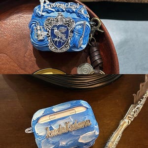 May include: Two blue and silver Airpod cases with different designs. One case features the Ravenclaw house crest with the words "Ravenclaw" in silver lettering. The other case has a blue and white marbled design with the words "Avada Kedavra" in silver lettering.