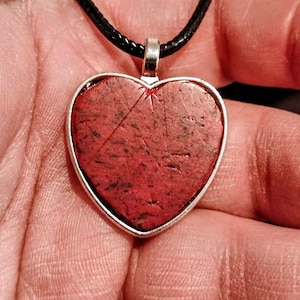 May include: A heart-shaped pendant featuring a textured, speckled red stone set in a silver-toned metal frame. The pendant is attached to a black cord necklace, showcasing a natural, porous surface with dark inclusions and a small etched starburst.