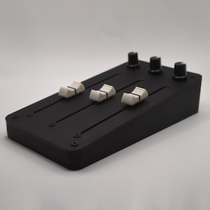 May include: A black audio mixer featuring three white sliders and three black knobs. The rectangular device has rounded edges and sits against a plain white backdrop. The sliders are positioned along the top surface.