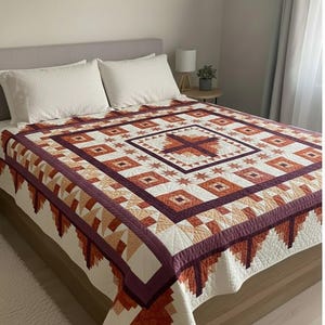 Puede incluir: Un edredón artesanal con un patrón geométrico en tonos crema, naranja y burdeos. El edredón presenta un diseño cuadrado central con un motivo de cruz, rodeado de formas repetidas de diamantes y estrellas. El edredón se exhibe en una cama con almohadas blancas.