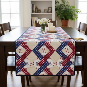May include: A patriotic table runner with a geometric pattern of red, white, and blue stars and stripes. The runner is placed on a dark wooden table, with a vase of white flowers and candles. The runner is approximately 60 inches long.