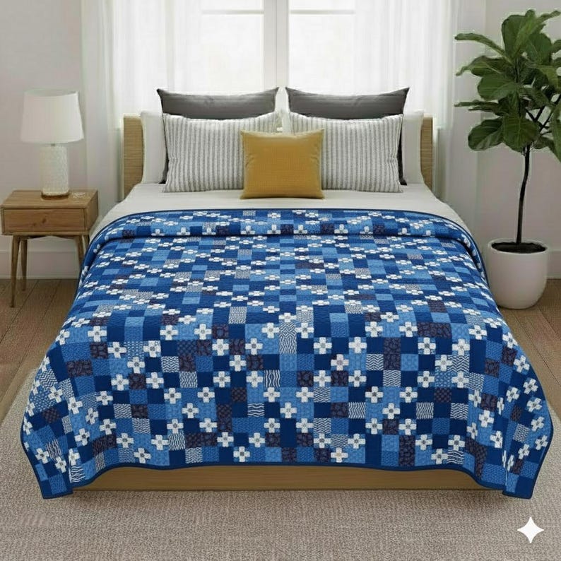 May include: A blue and white patterned quilt covers a bed. The quilt features a patchwork design with squares of varying shades of blue, white, and dark blue. The bed is made with white sheets and pillows, and a yellow pillow.