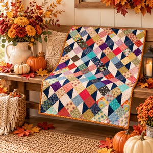 May include: A rectangular quilt featuring a patchwork design of various colorful floral and geometric fabric squares arranged in a repeating pattern, bordered by an orange trim, with a white background and dark blue triangular accents throughout.