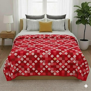 Op de afbeelding: Een rood-wit patroon quilt bedekt een bed, met een herhalend vierkant ontwerp en bloemaccenten. De quilt is het middelpunt, aangevuld met kussens en een neutraal gekleurd tapijt. Het bed staat voor een raam.