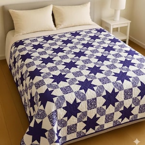 May include: A bed with a white comforter and two cream-colored pillows. The bedspread is a quilt with a repeating pattern of navy blue stars and white squares. A white nightstand with a lamp is visible in the background.
