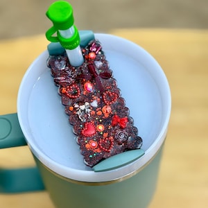 May include: A green travel mug with a decorative lid featuring red hearts, bows, and glitter. The lid has a green straw with the word "house" printed on it. The mug is green with a matching handle.