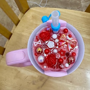 May include: A pink water bottle with a decorative top filled with red roses, pearls, and a gold bow. The bottle has a blue spout and a pink handle, perfect for carrying.