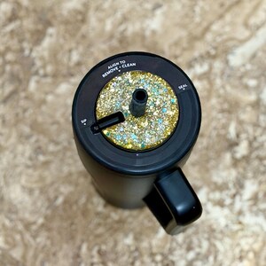 May include: Black travel mug with a handle, featuring a glittery gold and teal accent on the lid. The lid has text that reads "Align to Remove + Clean" and "Seal". The mug is designed for easy sipping.