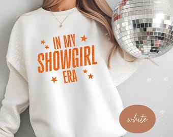 May include: White sweatshirt with the text "IN MY SHOWGIRL ERA" in orange glitter lettering. The design includes orange star accents. A disco ball is held in the background.