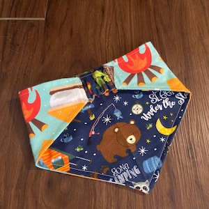 Puede incluir: Un bandana azul, naranja y marrón con temática de camping. El bandana presenta un oso, una fogata, una tienda de campaña y el texto "Gone Camping" y "Sleep Under the Stars".